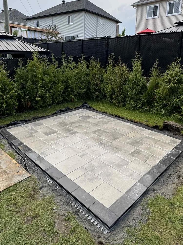 Patio en dalles de béton gris terminé, bordé de pavé foncé et de cèdres.